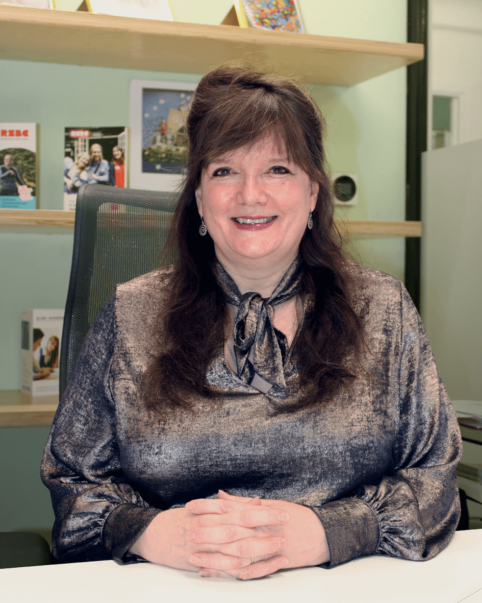 Julie, a light skinned woman with long brown hair sits at a desk with her hands folded in front of her. There are wooden shelves in the background. She is smiling broadly and wearing a metallic bronze coloured top.