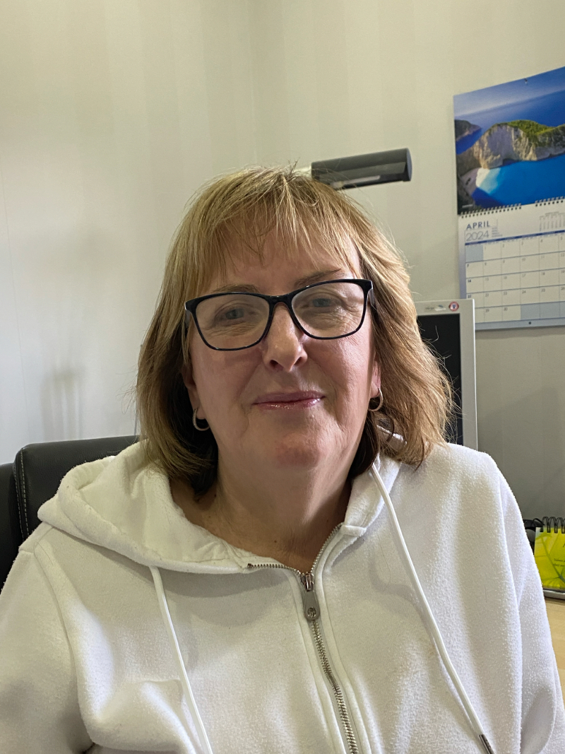 Sue, a light skinned woman with shoulder length blonde hair, wears glasses and a white hoody. She sits with her back to a desk and has a serious expression.