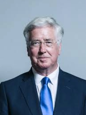 Michael, a light skinned man with grey hair wears metal glasses and a dark blue suit with a white shirt and light blue tie. He looks directly at the camera with a serious expression.