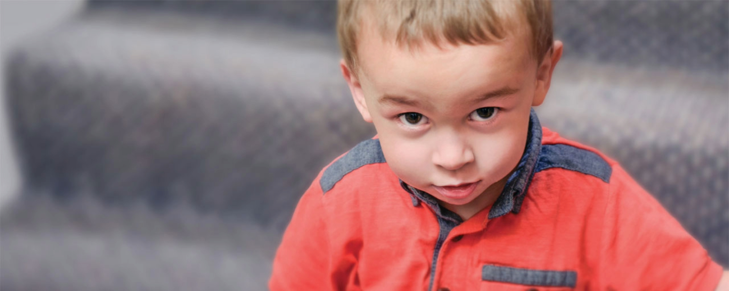 A light-skinned little boy with light brown hair sat on some stairs. He is looking up toward the camera with a cheeky half-grin.