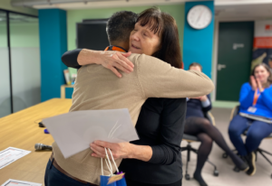 An elderly female volunteer hugging a male staff member in gratitude as he hands her a certificate and giftbag at an awards ceremony. People in the background are clapping.