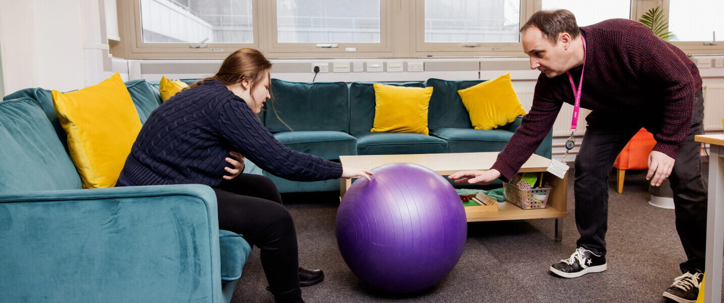 A light skinned Dorton College staff member wearing brown clothes and trainers rolls a large purple exercise ball towards a light skinned student with tied back brown hair, who's stretching towards it while she sits on a blue sofa.