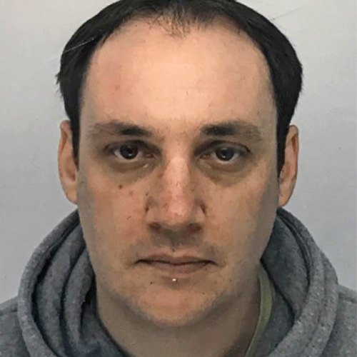 Alex, a light skinned man with receding dark hair and dark eyes, looks seriously into the camera. He is in front of a light grey background and wears a light grey hoodie.