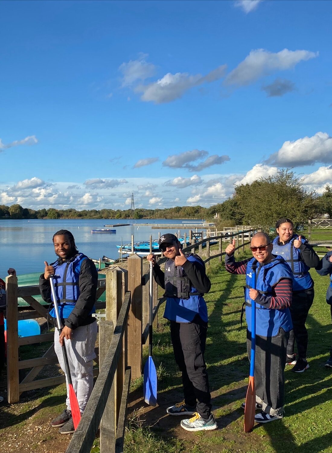 A group of vision impaired young people on a residential trip. They are standing in front of a lake, wearing life-vests and holding ores.