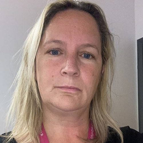 Chantelle is light skin toned with long straight blonde hair and blue eyes. She is standing against a white background and looking intently into the camera. She wears a black top and a pink lanyard is around her neck.