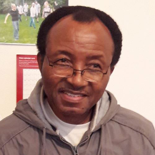 Charles, a dark skinned man with receding black hair, wears glasses and a grey hoodie with a white T shirt underneath. He is looking into the distance away from the camera and has a slight smile.