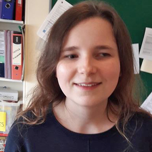 Charlotte, a light skinned woman with long dark hair, stands in front of a wall with folders and notices on it. She has a smile on her face and wears a dark coloured t shirt, looking slightly away from the camera.