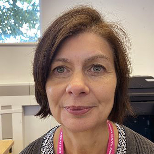 Colette, who has a light skin tone, blue eyes and short brown hair, sitting in an office. She has a slight smile on her lips and wears a grey cardigan and a black and white patterned top. She has a pink lanyard around her neck.