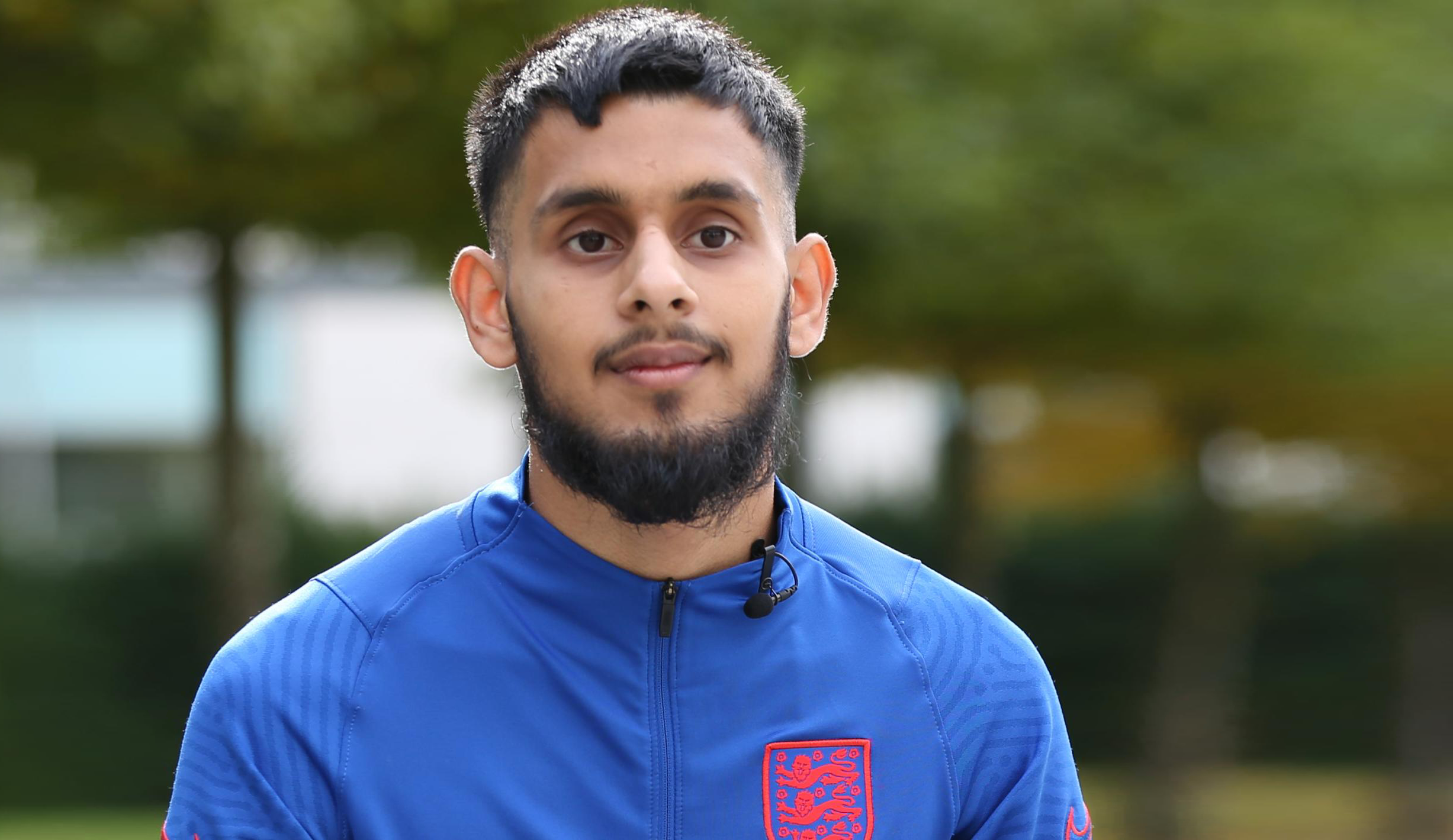 A medium-skinned young man with black hair and a black beard looks into the distance with a serious expression. He wears a blue sports top that has the three lions symbol in red on his chest.