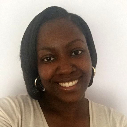 Genyne, a dark skinned woman with black hair and brown eyes, smiles happily at the camera. She wears large silver hooped earrings and a cream coloured top and is in front of a white background.