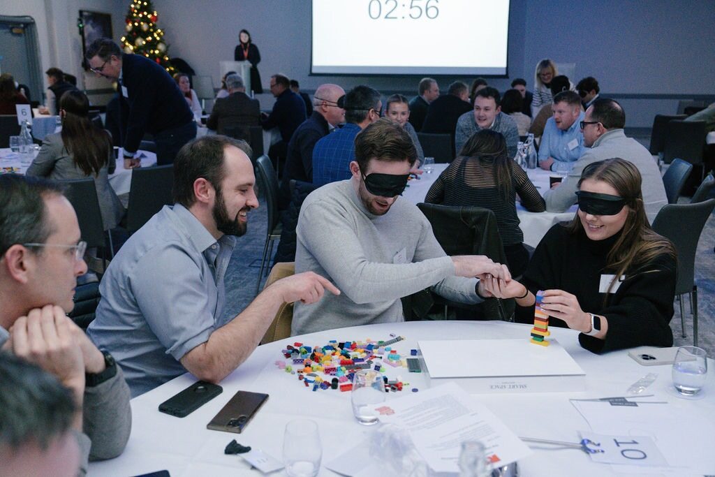 Three team members from Gresham House engaged in a team-building exercise using lego blocks at a conference table. Two team members are blind folded, while the other team member tries to help.