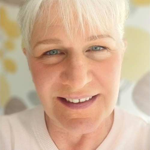 Hayley, a light skinned woman with light blonde hair and a fringe and light coloured eyes, wears a white top and smiles into the camera. The background is yellow and beige patterns on white.