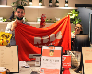 Two people standing behind the counter of a cafe, holding an RSBC banner.