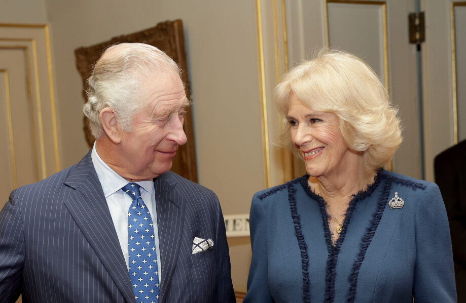 King Charles III, a light skinned man with grey hair, wears a grey suit and blue tie with white dots. He stands next to at Queen Camilla, a light skinned woman with blonde hair, and they are smiling at each other. She wears a mid blue dress with dark blue piping.