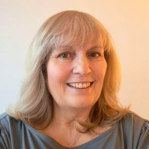 Lynne, a light skinned woman with long blonde hair and a fringe, smiles broadly as she looks into the camera. The background is a light beige colour and she wears a grey top.