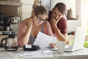 A young male and female couple in their kitchen at a table looking over a piece of paper with a breakdown of their income and expenditure. The lady is sat at the desk with her hand on her neck, leaning on the desk and the male is behind her, leaning on the desk with his elbow and hand to his face. Both look concerned.