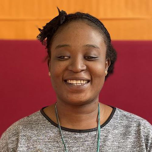 Monsurat, a dark skinned person with black curly hair swept to the back of her head, is smiling widely as she looks down slightly to the left hand side. She wears a grey top with black piping around the neck and stands in front of a background that is dark red at the bottom and orange at the top.