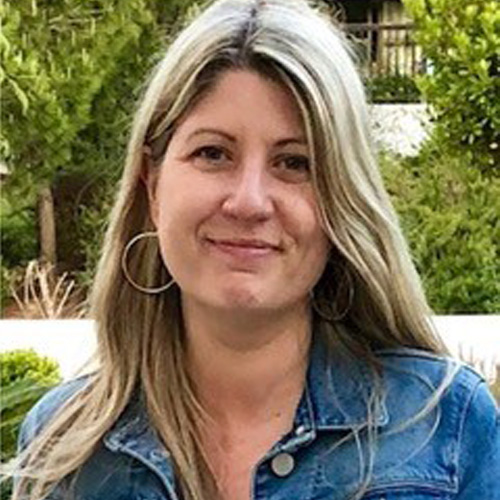 Sarah, a light skinned woman with long blonde hair, is smiling at the camera. She is standing in front of some trees and wears a blue denim jacket and hooped earrings.