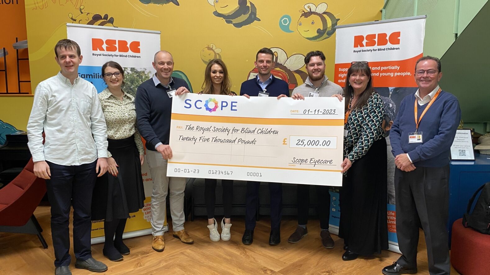 Group of eight people smiling and holding a large check for £25,000 from SCOPE Eyecare in front of RSBC banners.