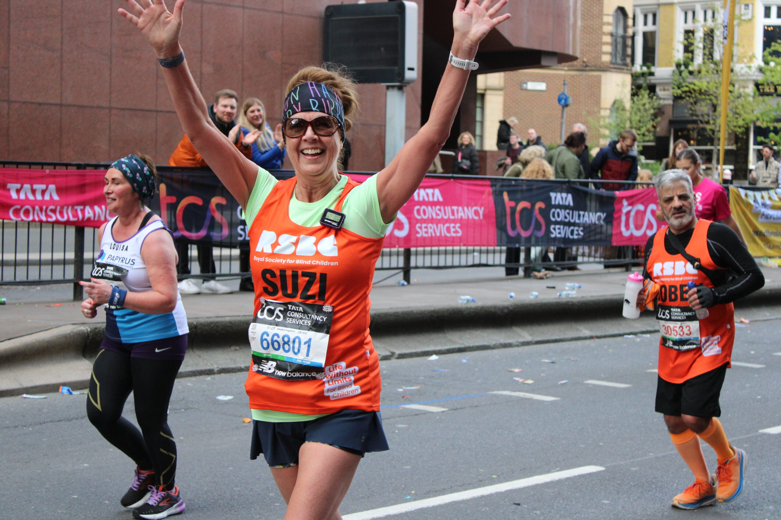 Two RSBC London Marathon runners wearing orange tabards are running as a pair, with the woman in front raising her arms and smiling happily as she leads her VI team-mate behind.