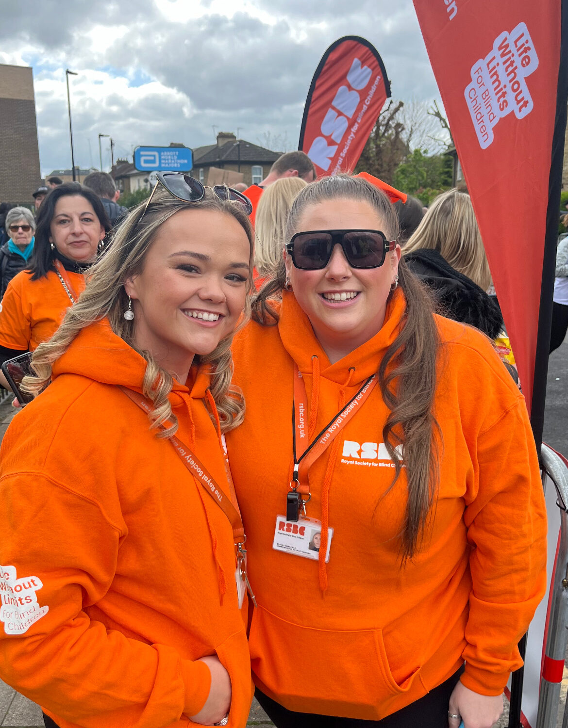 Two light skinned RSBC supporters standing outside and smiling broadly at the camera. They wear orange RSBC hoodies and lanyards.