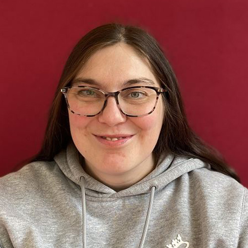 Zara, a light skin toned person with straight, long brown hair, wears tortoiseshell glasses and a happy smile as she looks into the camera. She's in a grey hooded top with a white logo on the left hand side, and stands in front of a deep red background.