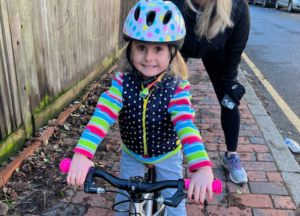 A 5-year-old girl in bright coloured striped jumper, pulka dot waistcoat and colourful cycle helmet, sitting on her bicycle on the pavement. Her mum is behind her.