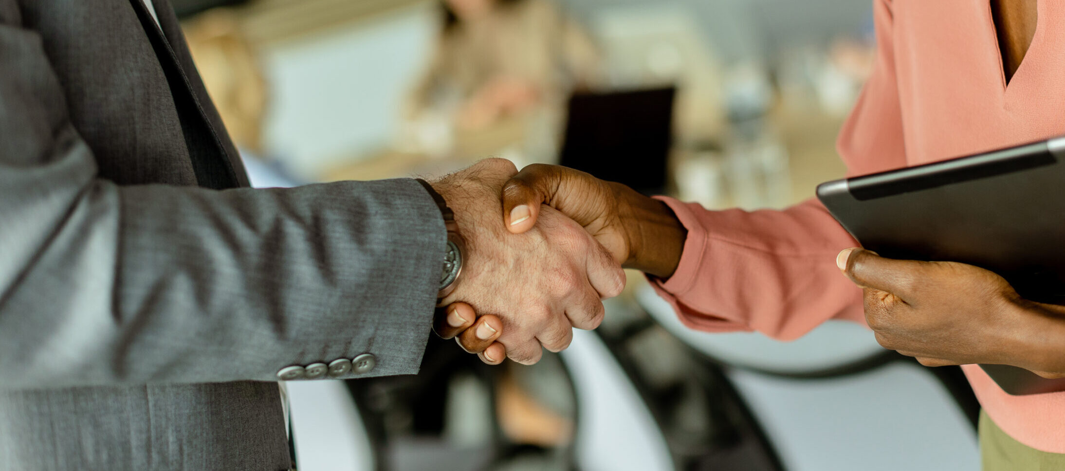 Two people's arms, one with a light skin tone and grey clothing and one with a medium dark skin tone and pink clothing, shake hands, with an office meeting room in the background.