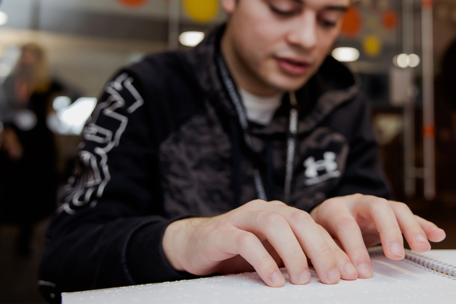 A medium light skinned Dorton College student wears a black top as he sits and looks downwards, reading a braille book.