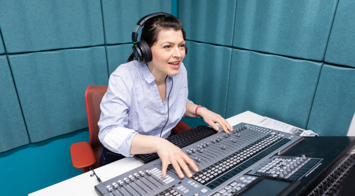 A light-skinned woman with dark brunette hair using the RSBC media suite to record a podcast. She is sat at a recording desk with a large sound mixing board, wearing headphones.