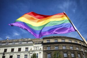 An LGBTQIA+ Pride flag waving against blue sky and buildings.