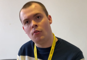 A young man wearing a black top and yellow lanyard. He is about to speak.