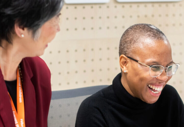 A light skinned woman with grey hair and a dark red top is facing away from the camera, talking to her colleague who has medium dark skin, closely cropped hair and metal glasses. They are smiling happily.