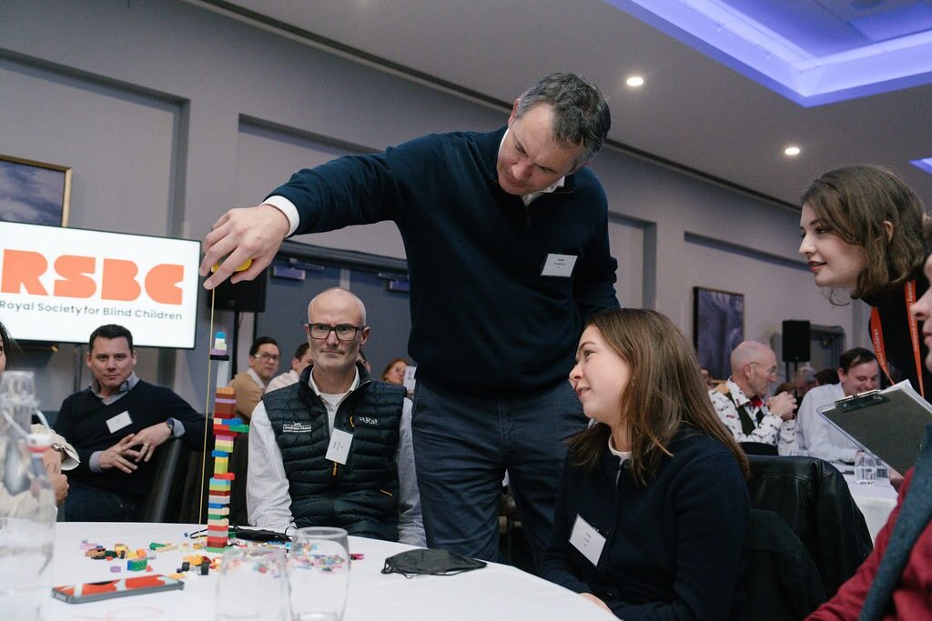 Three team members from Gresham House taking part in a team-building exercise using lego blocks at a conference table. There is a small lego tower built on the table and another team member is measuring the tower.