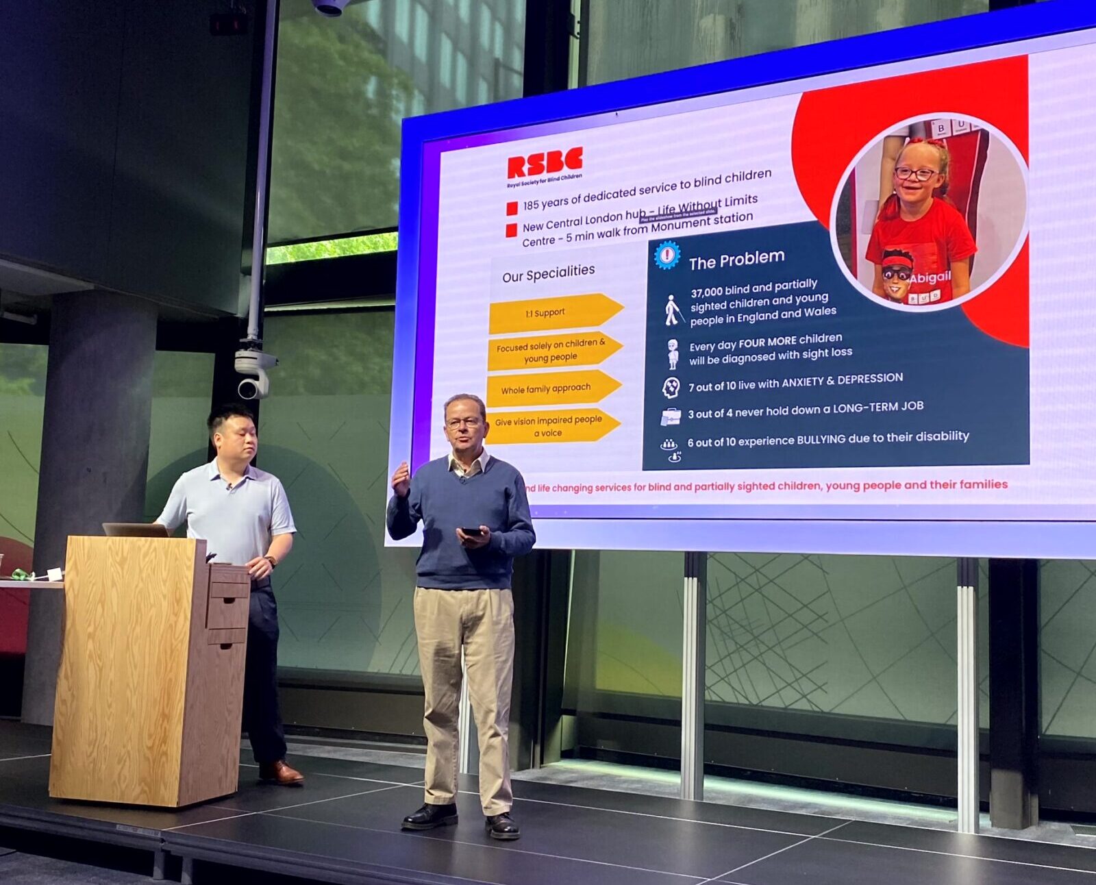 Two RSBC team members stand on stage delivering a presentation. One is at a wooden lectern, while the other stands to the right of them in front or a large screen and speaks to the audience.