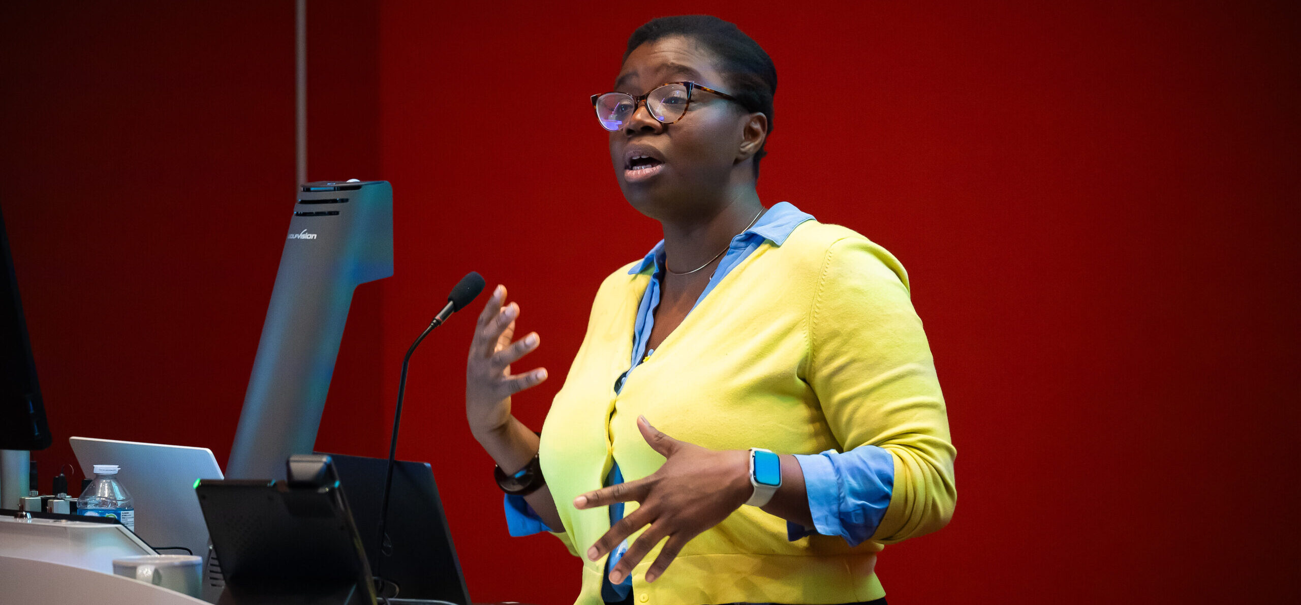 A dark-skinned woman with glasses and dark hair wears a yellow cardigan and bright blue shirt. She stands behind a lectern, speaking into a microphone.