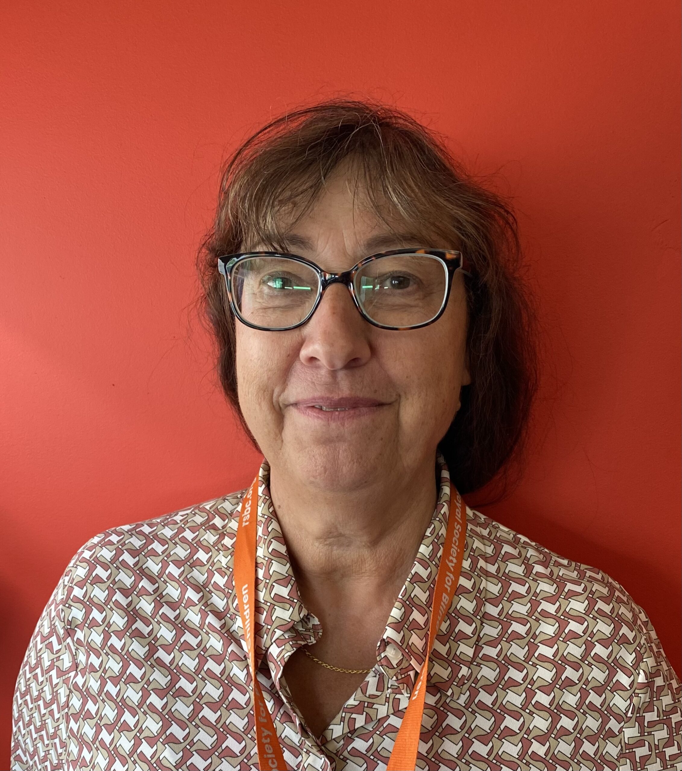 Frederique, a light skinned woman with glasses and brown eyes, gives a slight smile as she looks into the camera. She wears a beige and white patterned top and an orange RSBC lanyard as she stands against an orange wall.