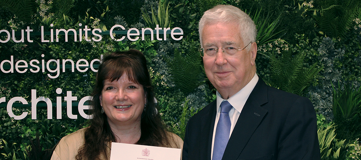 Julie, a light skinned woman with long dark hair stands next to Michael, a light skinned man with grey hair who wears glasses and a dark suit with blue tie. She holds a letter up to the camera. Both are smiling, and they're standing in front of some green foliage and white lettering.