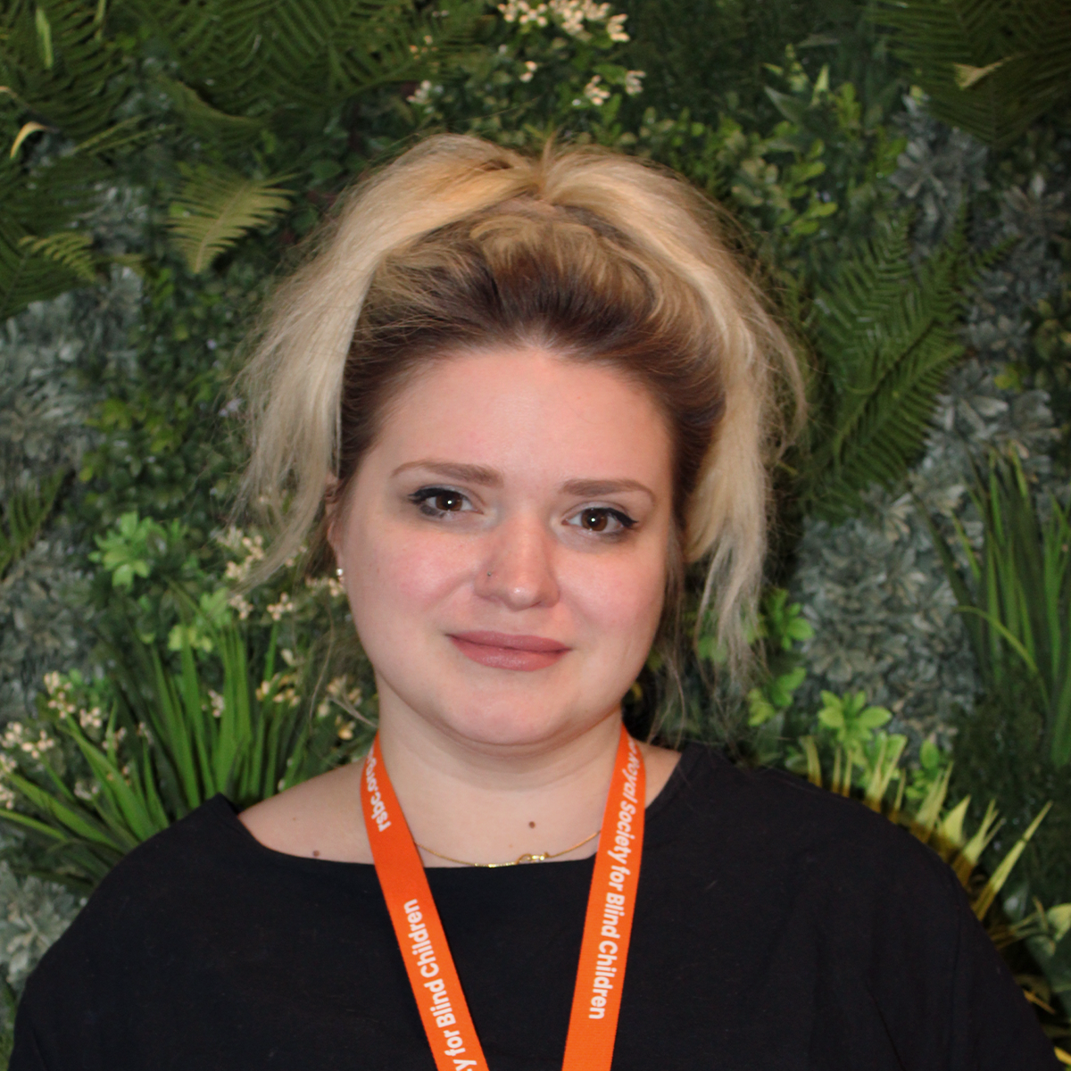 Lily, a light skinned woman with blonde hair swept up on top of her head gives a slight smile to the camera as she stands in front of a wall of green vegetation. She wears a black round neck top and an orange RSBC lanyard.