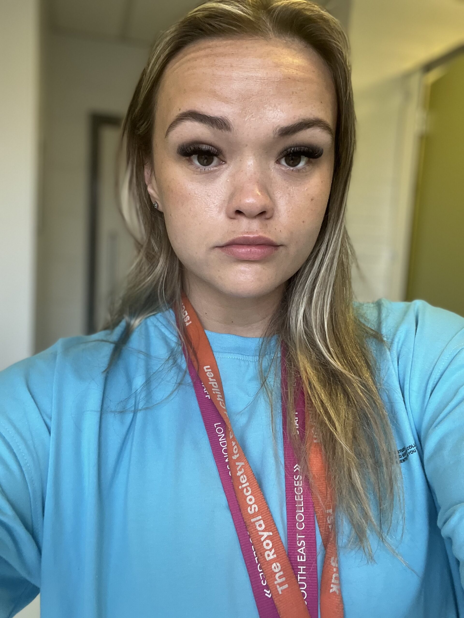 Michaela, who has a light skin tone, long blonde hair and wide dark eyes, stares intently into the camera. She is wearing a turquoise top and two lanyards round her neck, one pink, one orange.