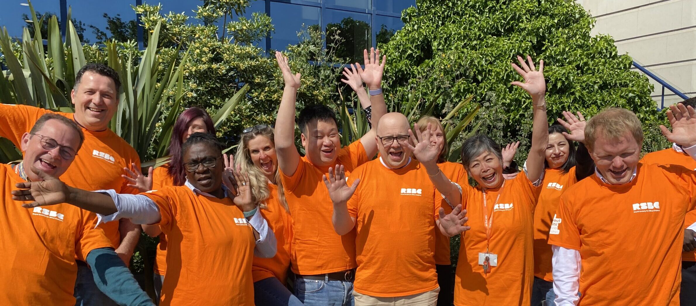 A group photo of RSBC staff members standing outside in front of some greenery with blue sky above them. They're all wearing orange t-shirts with an RSBC logo in white, while smiling and waving at the camera.