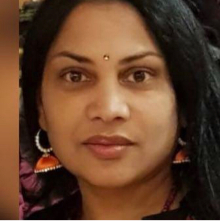 Srilatha has a medium skin tone and long, straight black hair and brown eyes. She stares directly into the camera, looking serious. She is wearing large orange earrings and has a single bindi between her eyebrows.