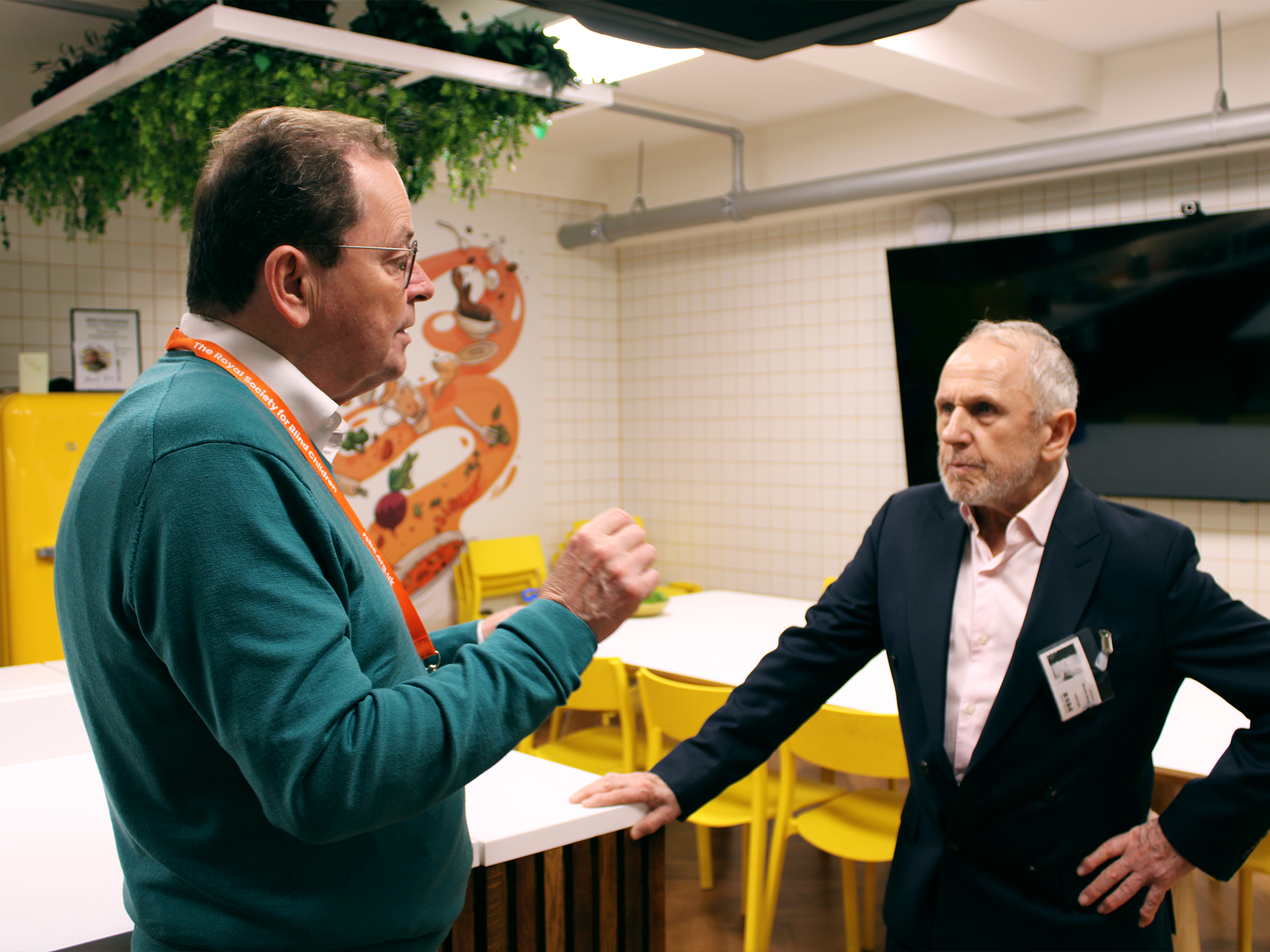 An RSBC staff member in a green jumper stands in front of a white table with yellow chairs, his hands raised as he talks to RSBC Ambassador Wayne Sleep. Wayne has his hand resting on a white table, wears a dark jacket and light open necked shirt, and is listening to the member of staff.