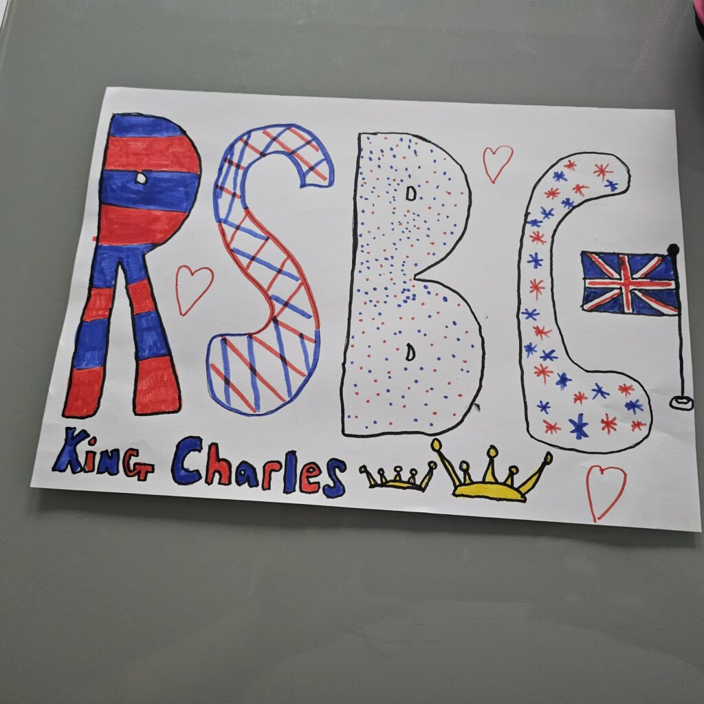 A felt tipped drawing in red, white and blue of the enlarged letters "RSBC' with a flag, 2 yellow crowns and hearts, with the name "King Charles" at the bottom of the letters 