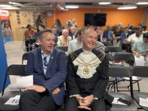 Two gentlemen sat at the front of the audience at a prizegiving ceremony. One of them is dressed like a Lord mayor. 