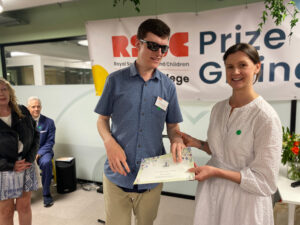 A lady giving a student, with dark glasses, his certificate at the front of a prizegiving ceremony.