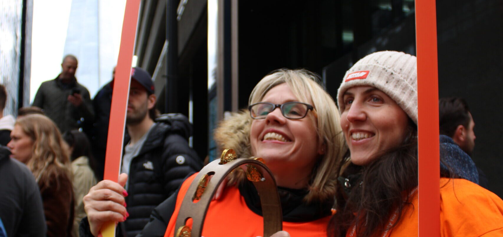 Two RSBC volunteers - the one on the left is a light-skinned woman with blonde hair and glasses. She's holding a tambourine, and the one on the right is a light-skinned woman with long brown hair who's wearing a beanie. Both volunteers are wearing orange RSBC t-shirts and holding a novelty photo frame.