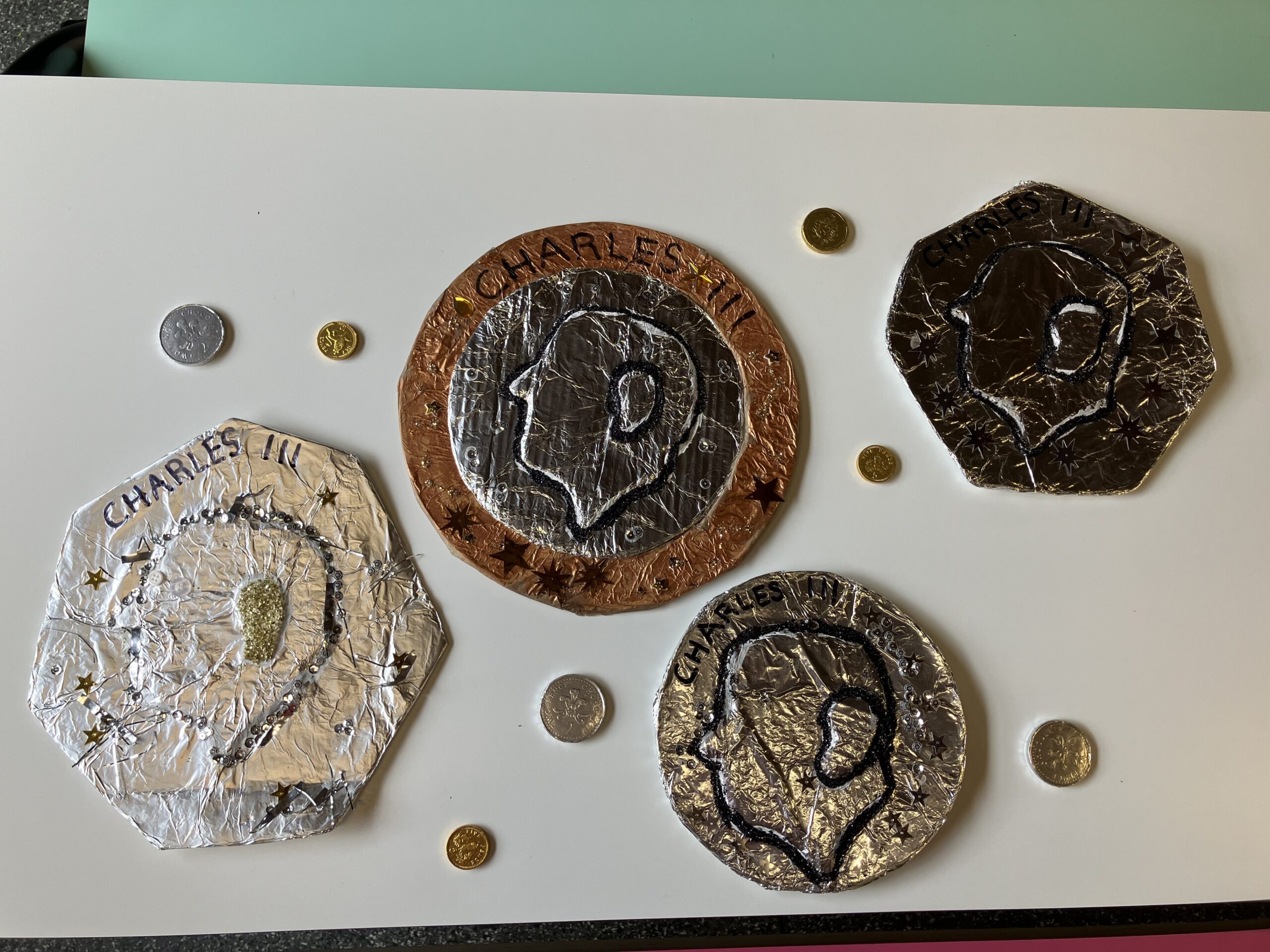 Four enlarged sculptured coins with King Charles' face, alongside normal sized coins on a white table