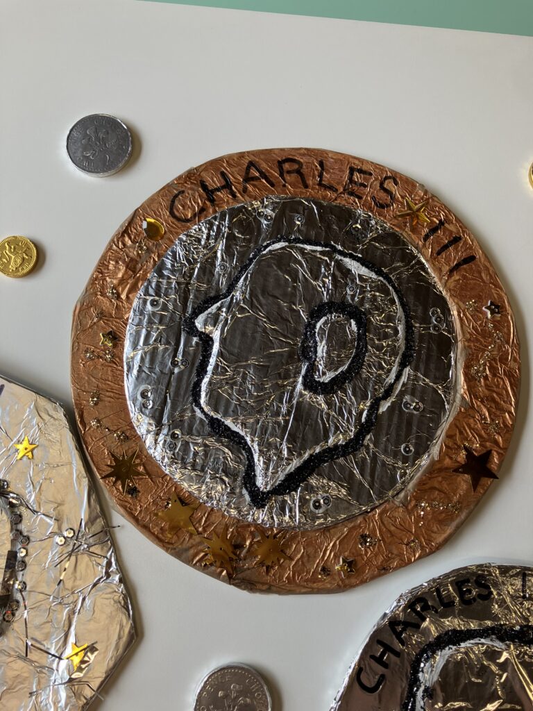 An enlarged sculptured coin with King Charles' face, alongside normal sized coins on a white table