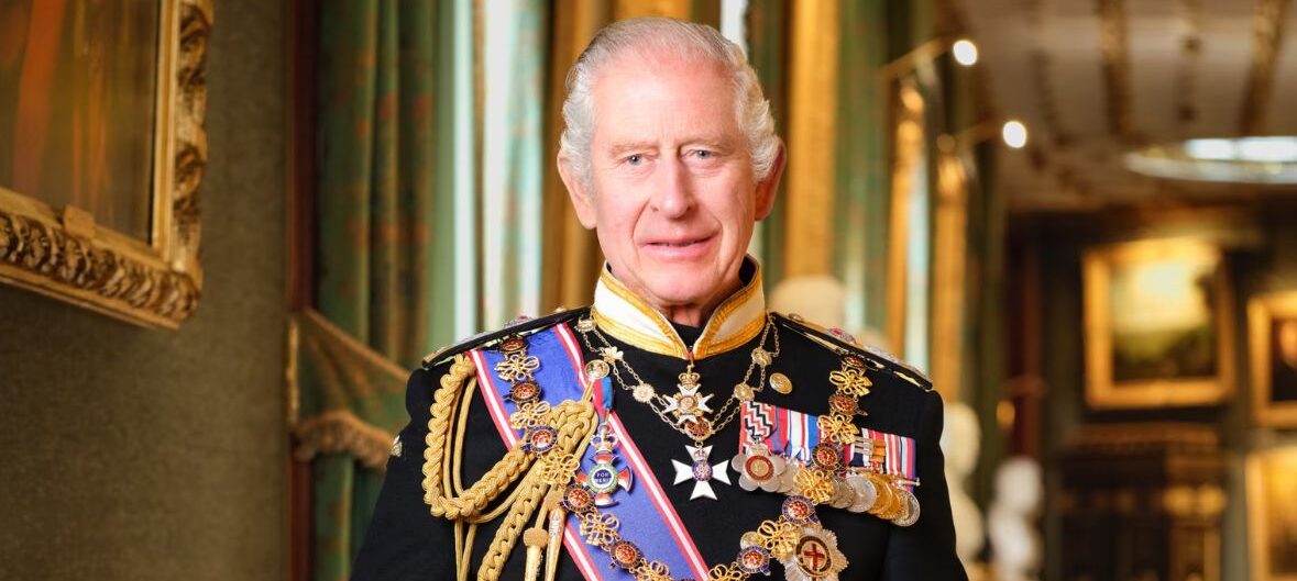 King Charles III, a light-skinned older man with grey hair. He smiles slightly as he wears a ceremonial military uniform, adorned with medals. He stands in a room with pictures on the walls, and is smiling slightly.